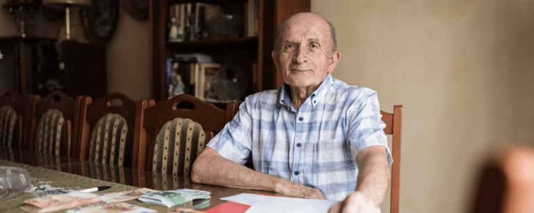 Older man sitting at a dining table with papers and euro banknotes spread out in front of him, looking directly at the camera in a home environment.