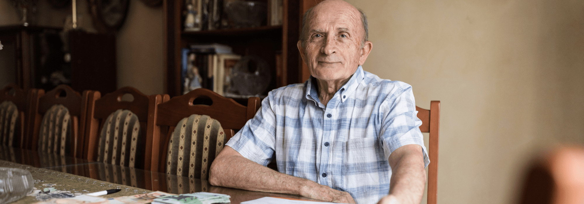 Older man sitting at a dining table with papers and euro banknotes spread out in front of him, looking directly at the camera in a home environment.