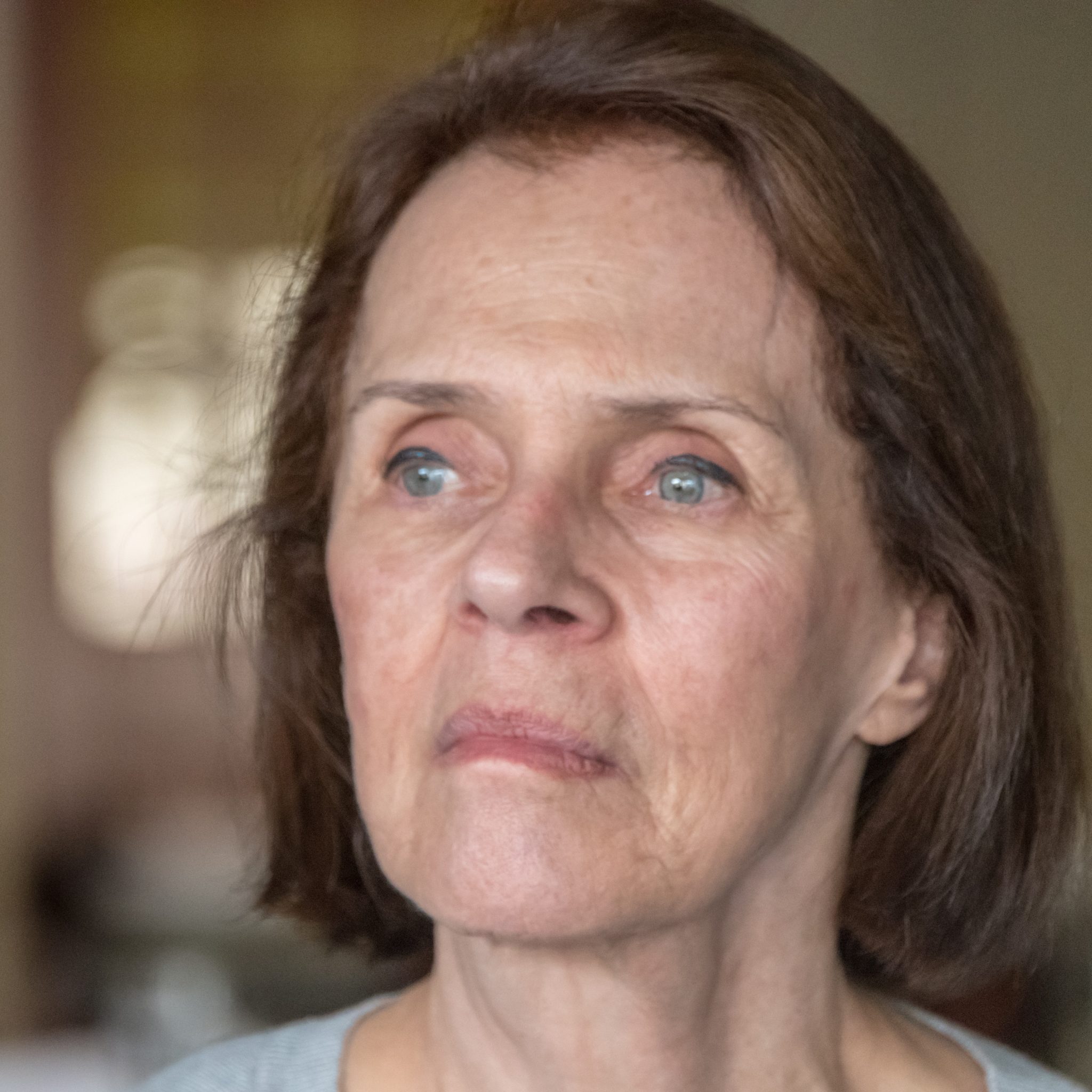 Close-up portrait of an older woman with short brown hair looking ahead with a reflective, emotional expression in a softly lit indoor setting.