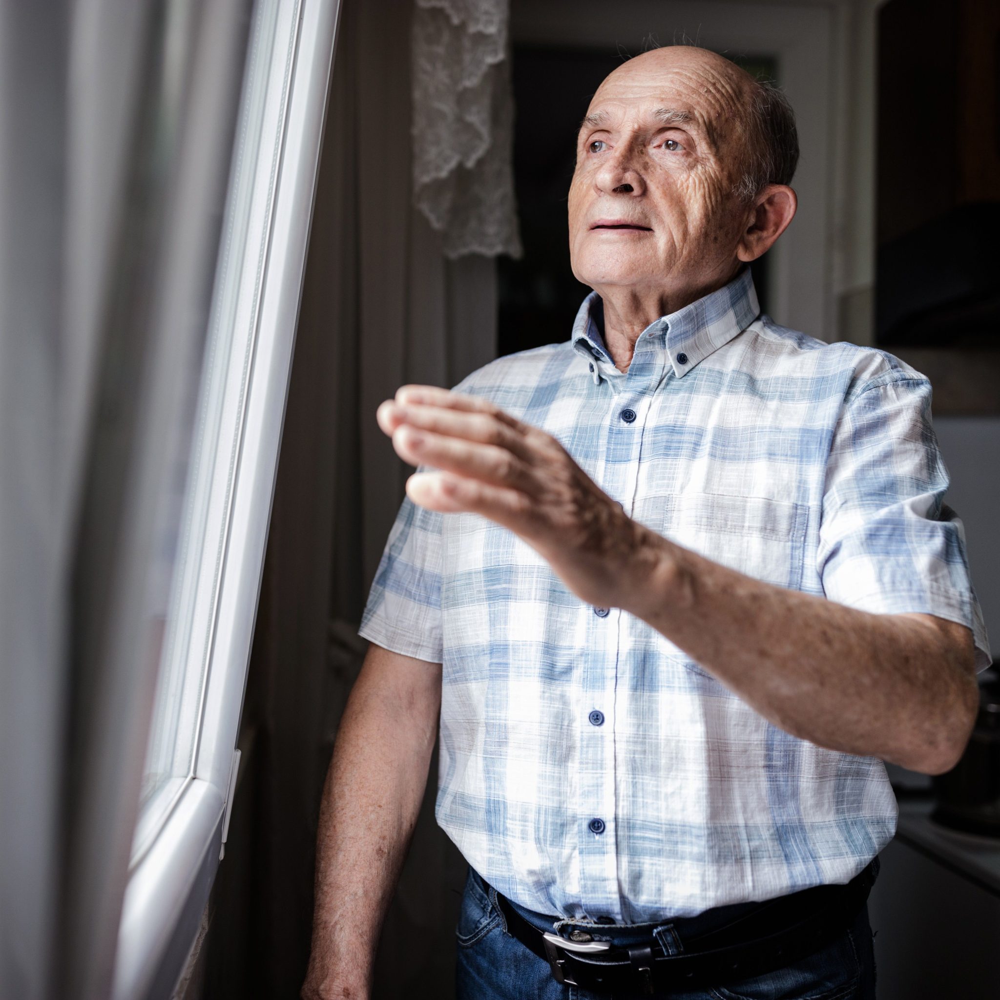 Older man standing beside a window in a kitchen, raising one hand slightly as he looks outside with a thoughtful expression.